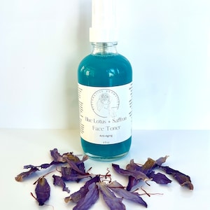 May include: A blue glass bottle with a white spray top and a white label that reads "Blue Lotus + Saffron Face Toner Anti-Aging 4 fl oz".  Dried purple flower petals are scattered on a white surface.