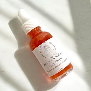 Może przedstawiać: Przezroczysta szklana butelka z pomarańczowym płynem w środku. Butelka ma białą etykietę z czarnym tekstem, kt&oacute;ry brzmi "Renew + Resurface Face Drops Glycolic Acid + Peptides + Niacinamide 1 fl oz".