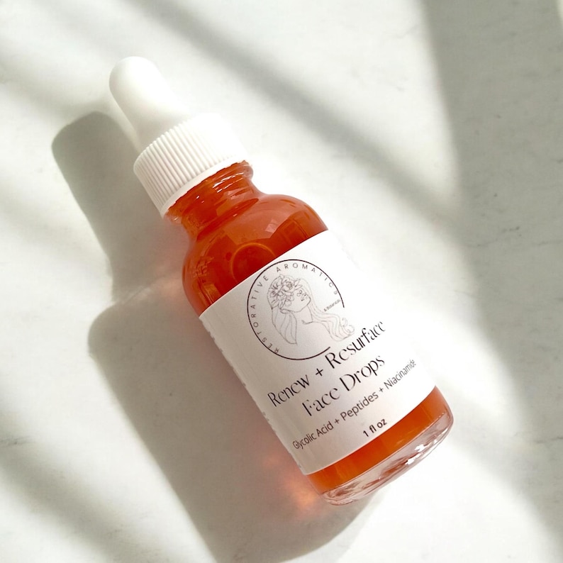 Może przedstawiać: Przezroczysta szklana butelka z pomarańczowym płynem w środku. Butelka ma białą etykietę z czarnym tekstem, kt&oacute;ry brzmi "Renew + Resurface Face Drops Glycolic Acid + Peptides + Niacinamide 1 fl oz".