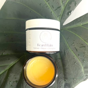 May include: A small glass jar with a yellow beard balm inside. The jar has a white label that reads "Beard Balm" and "Hydrate + Repair + Strengthen" with a small logo. The jar is sitting on a green leaf.