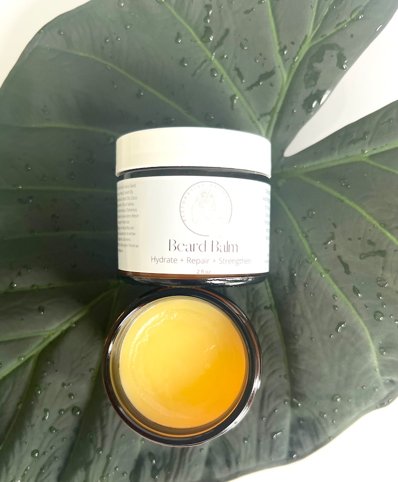May include: A small glass jar with a yellow beard balm inside. The jar has a white label that reads "Beard Balm" and "Hydrate + Repair + Strengthen" with a small logo. The jar is sitting on a green leaf.