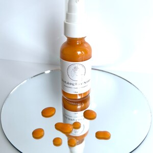 May include: A small amber glass bottle with a white pump top dispenser. The bottle is labeled "Revitalizing Face Serum" with the ingredients listed as "Astaxanthin + CoQ + Vitamin C". The bottle is sitting on a silver round mirror surface with small drops of the serum scattered around it.