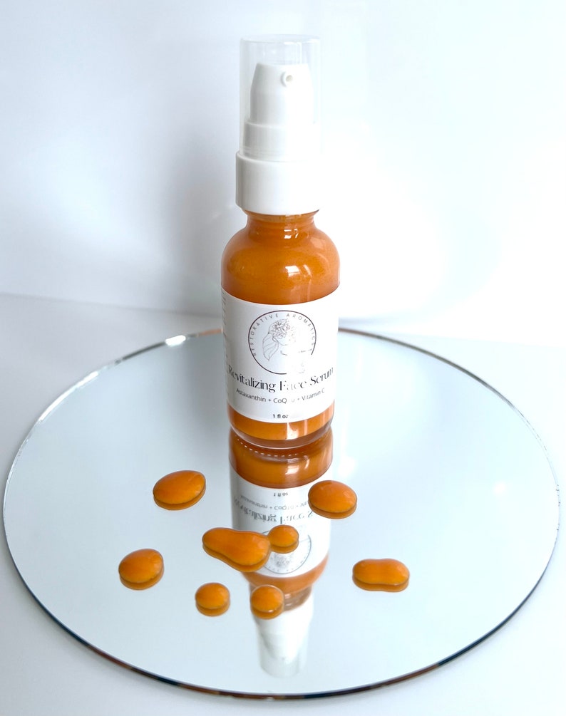 May include: A small amber glass bottle with a white pump top dispenser. The bottle is labeled "Revitalizing Face Serum" with the ingredients listed as "Astaxanthin + CoQ + Vitamin C". The bottle is sitting on a silver round mirror surface with small drops of the serum scattered around it.