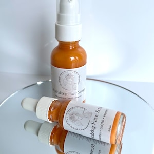 May include: Two amber glass bottles with dropper tops filled with a golden liquid. The bottles are labeled "Revitalizing Face Serum" with the ingredients listed below. The bottles are sitting on a reflective surface.