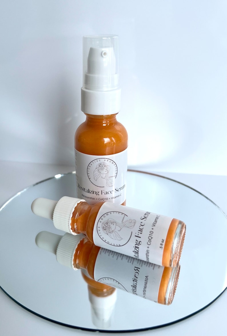 May include: Two amber glass bottles with dropper tops filled with a golden liquid. The bottles are labeled "Revitalizing Face Serum" with the ingredients listed below. The bottles are sitting on a reflective surface.
