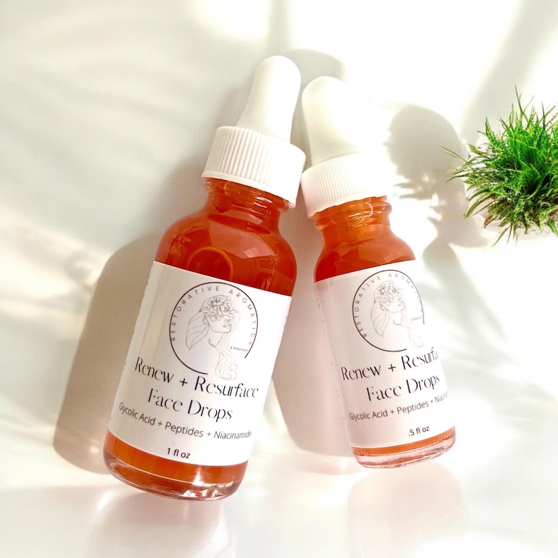 Może przedstawiać: Dwie bursztynowe butelki szklane z białymi zakraplaczami i białymi etykietami. Etykiety mają kwiatowy wz&oacute;r i tekst "Renew + Resurface Face Drops". Mniejsza butelka ma napis "5 fl oz", a większa "1 fl oz".