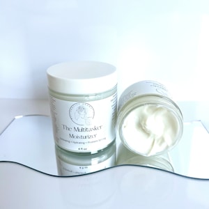 Può includere: Un barattolo bianco di crema idratante multifunzione "The Multitasker Moisturizer". Il barattolo è etichettato con il nome del prodotto, "Exfoliating + Hydrating + Problem Solving", e la dimensione, "4 fl oz".