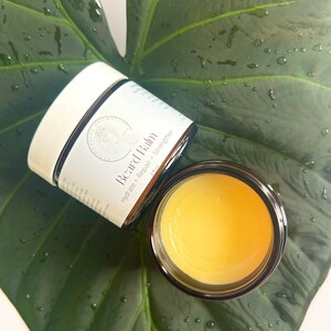 May include: A glass jar of beard balm with a white lid. The jar is labeled "Beard Balm" and "Hydrate + Repair + Strengthen". The jar is sitting on a green leaf.
