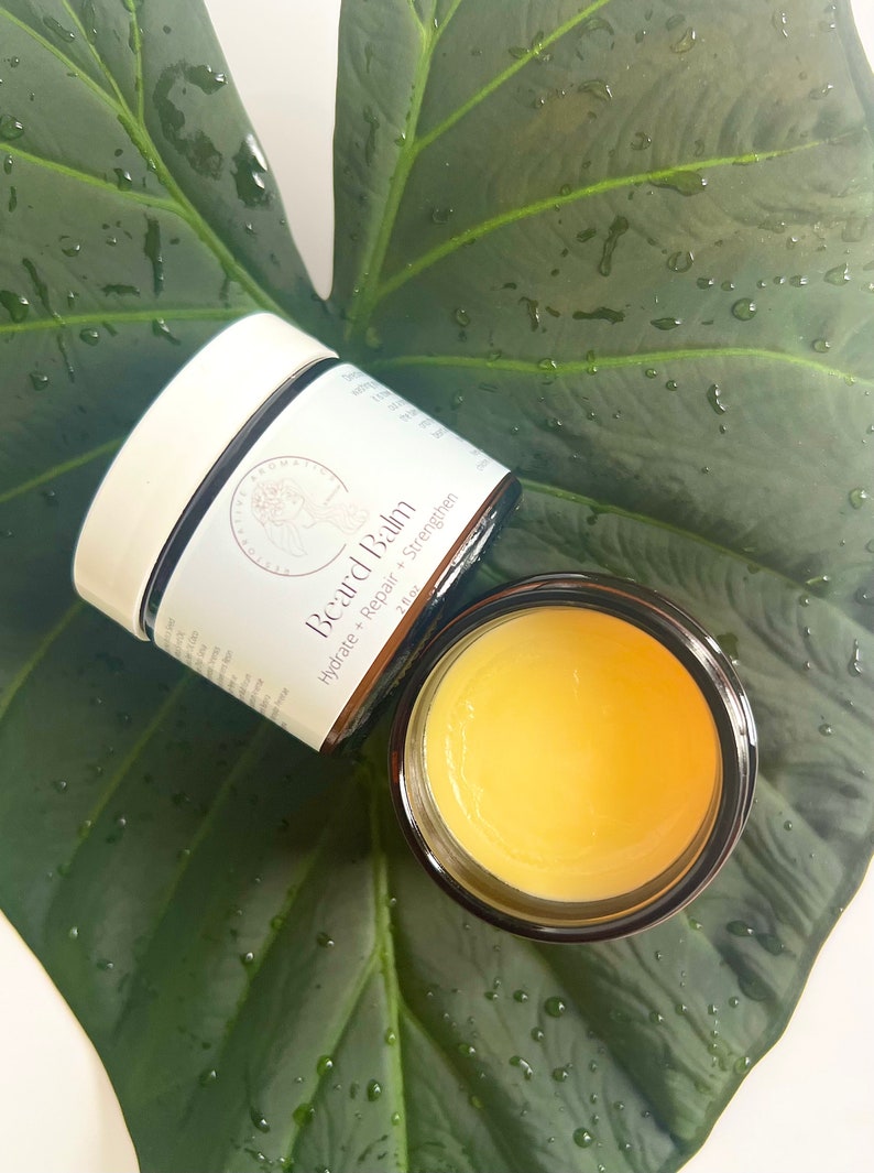 May include: A glass jar of beard balm with a white lid. The jar is labeled "Beard Balm" and "Hydrate + Repair + Strengthen". The jar is sitting on a green leaf.