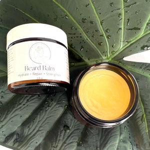 May include: A small jar of beard balm with a brown lid and a white label that says "Beard Balm" and "Hydrate + Repair + Strengthen" on a green leaf background.