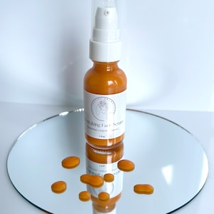 May include: A clear glass bottle with a white pump top filled with a golden liquid. The bottle is labeled "Vitalizing Face Scrub" with the ingredients "Adansonia + CoQ10 + Vitamin C" listed below. The bottle is sitting on a round, silver-colored mirror with several golden drops of liquid scattered around it.