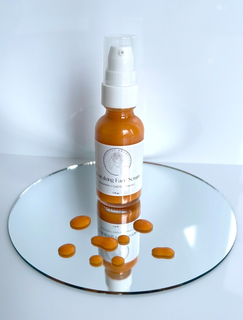 May include: A clear glass bottle with a white pump top filled with a golden liquid. The bottle is labeled "Vitalizing Face Scrub" with the ingredients "Adansonia + CoQ10 + Vitamin C" listed below. The bottle is sitting on a round, silver-colored mirror with several golden drops of liquid scattered around it.