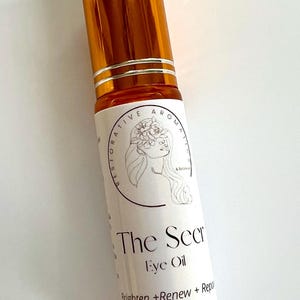 May include: A small, amber-colored glass bottle with a gold cap and a white label. The label reads "The Secret Eye Oil" and "Brighten + Renew + Repair". The bottle contains 10 ml of oil.