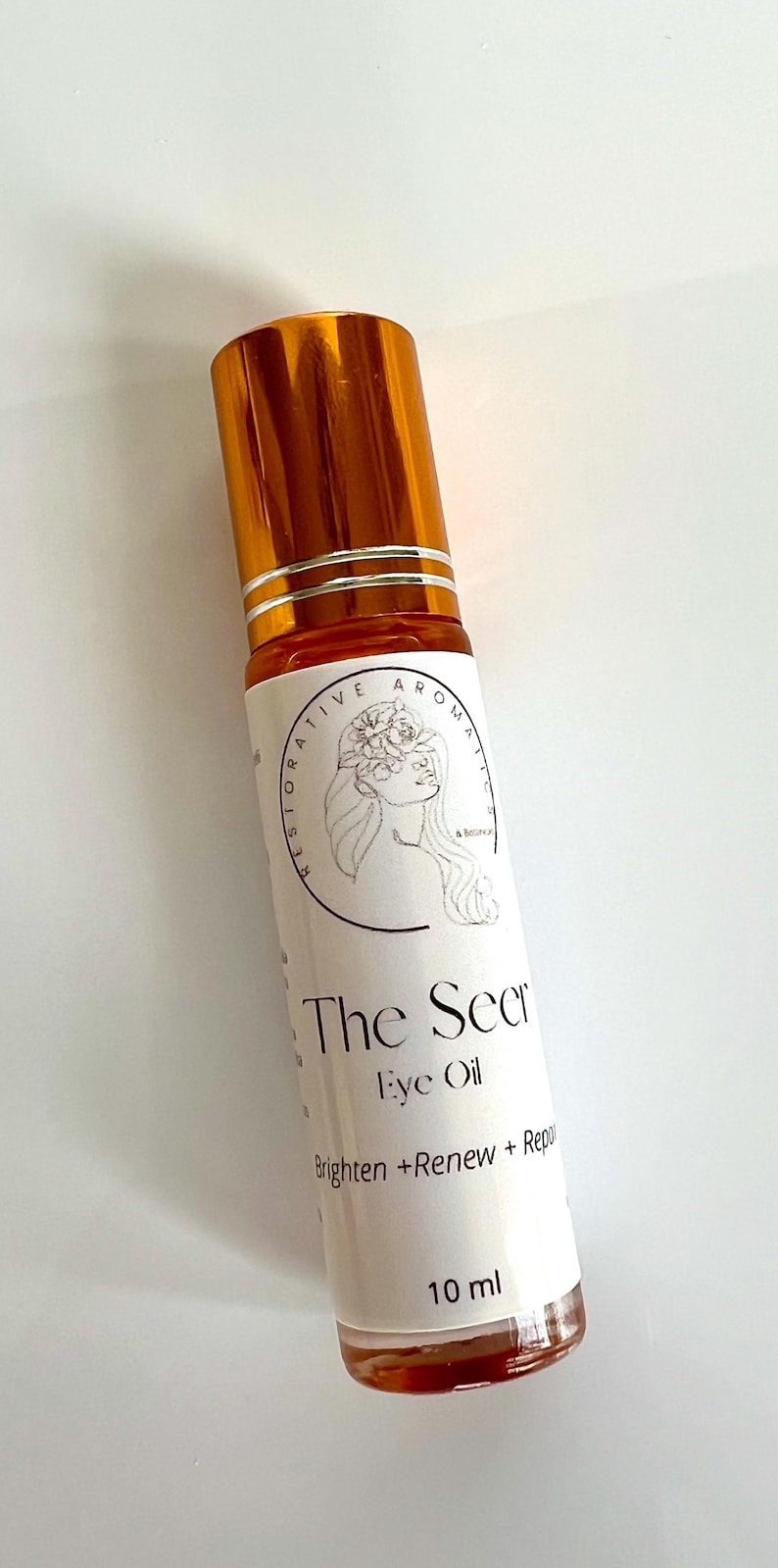 May include: A small, amber-colored glass bottle with a gold cap and a white label. The label reads "The Secret Eye Oil" and "Brighten + Renew + Repair". The bottle contains 10 ml of oil.