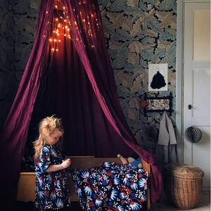 May include: A deep burgundy canopy bed with fairy lights, set against a floral wallpaper. A child in a patterned dress stands near the bed, which is covered with a blue, white, and red floral blanket. A wicker basket is also visible.