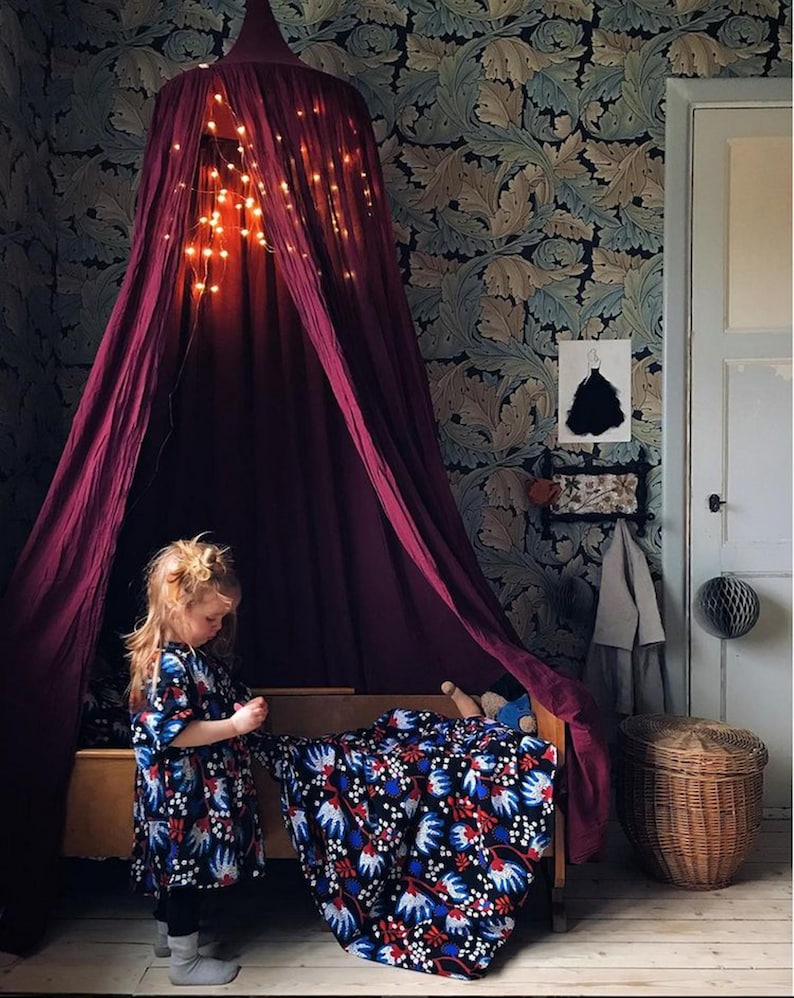 May include: A deep burgundy canopy bed with fairy lights, set against a floral wallpaper. A child in a patterned dress stands near the bed, which is covered with a blue, white, and red floral blanket. A wicker basket is also visible.