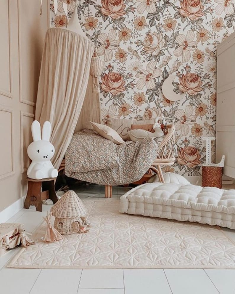May include: A child's bedroom with floral wallpaper and a patterned rug. Includes a white bunny lamp, a wicker chair, and a floor cushion. The room is decorated in soft, neutral tones, creating a warm and inviting atmosphere.