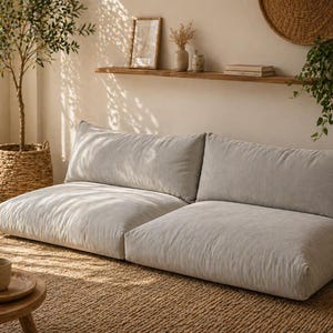 May include: A light grey, low-level sofa with two large cushions on a woven jute rug. A small wooden table with a cup and plate is in front of the sofa. A potted tree and decorative items complete the cosy, neutral-toned living space.