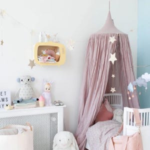 May include: A child's bedroom with a pink canopy over a cot. A yellow TV-shaped shelf displays toys. A plush monkey, doll, and bunny sit on a white cabinet. Star decorations and a mobile enhance the room's playful atmosphere.