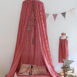 May include: A dusky pink canopy bed with draped fabric, a star-shaped mobile, and fairy lights. The bed has pillows and blankets. A dress form with a matching dress and a star decoration are nearby.