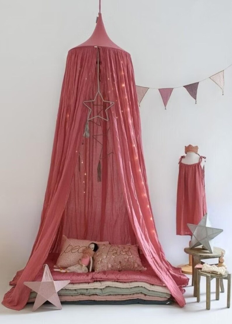 May include: A dusky pink canopy bed with draped fabric, a star-shaped mobile, and fairy lights. The bed has pillows and blankets. A dress form with a matching dress and a star decoration are nearby.