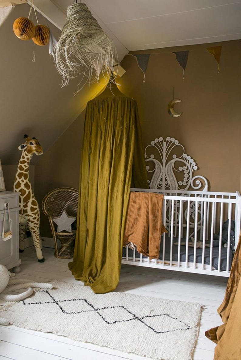 May include: A child's bedroom with a white cot, a giraffe toy, and a woven rug with a black geometric pattern. A mustard-coloured canopy hangs over the cot, and a star-shaped pillow sits on a wicker chair.