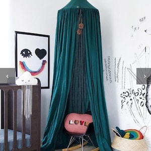 May include: A teal canopy hangs in a child's room, draped over a small chair with a pink pillow that says "LOVE". A dark wood cot and a framed print with a smiley face and rainbow are also in the room. A woven basket with a rainbow toy sits on the floor.