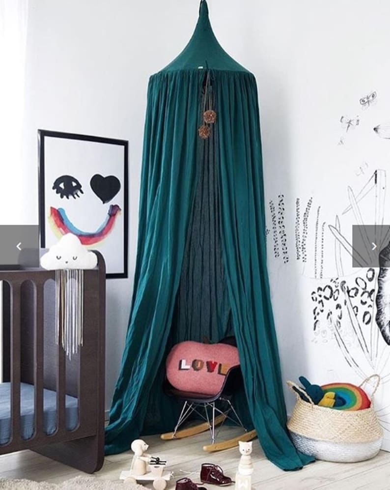 May include: A teal canopy hangs in a child's room, draped over a small chair with a pink pillow that says "LOVE". A dark wood cot and a framed print with a smiley face and rainbow are also in the room. A woven basket with a rainbow toy sits on the floor.