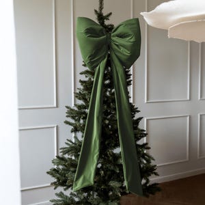 May include: A large, olive green velvet bow decorates a Christmas tree. The bow is centrally placed and features long, flowing tails. The tree stands against a white wall with rectangular panels, creating a festive holiday display.