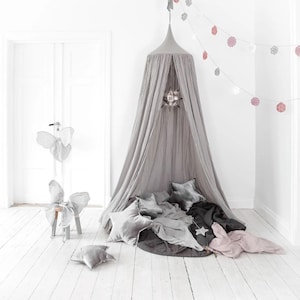 May include: A grey canopy bed with star-shaped cushions and blankets. A decorative star hangs inside the canopy. A garland of pink and white paper decorations hangs from the ceiling. A decorative chair with butterfly wings is in the background.