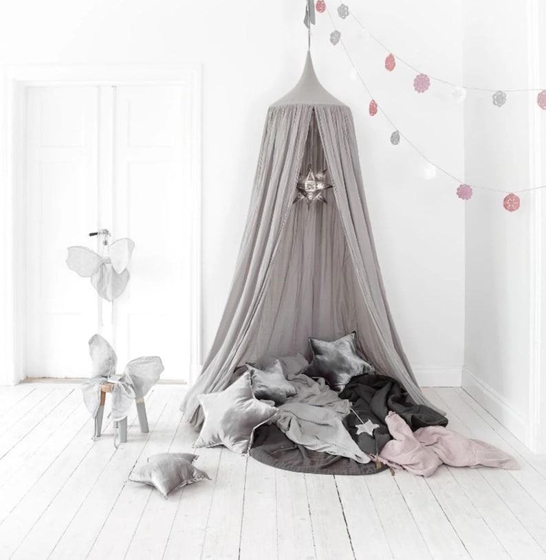 May include: A grey canopy bed with star-shaped cushions and blankets. A decorative star hangs inside the canopy. A garland of pink and white paper decorations hangs from the ceiling. A decorative chair with butterfly wings is in the background.