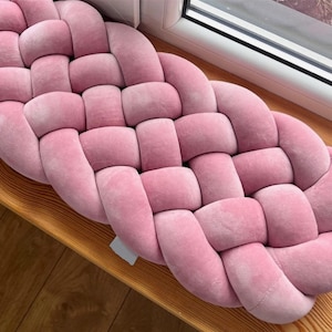 May include: A pink, braided cushion with a soft, velvety texture. The cushion is made of woven fabric and is designed in a decorative pattern. The cushion is placed on a wooden surface near a window.