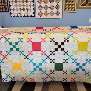 Può includere: Una trapunta fatta a mano con uno sfondo grigio chiaro e un motivo geometrico colorato. La trapunta presenta quadrati e forme a croce in vari colori, tra cui rosa, giallo, verde e blu. La trapunta è esposta su un telaio per trapuntatura.