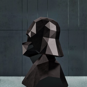 Puede incluir: Una escultura de busto geom&eacute;trico negro con un dise&ntilde;o facetado. La escultura tiene un acabado oscuro y mate y est&aacute; colocada sobre un fondo gris. El busto parece ser una representaci&oacute;n estilizada de una cabeza y hombros.