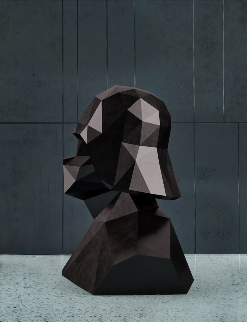 Puede incluir: Una escultura de busto geom&eacute;trico negro con un dise&ntilde;o facetado. La escultura tiene un acabado oscuro y mate y est&aacute; colocada sobre un fondo gris. El busto parece ser una representaci&oacute;n estilizada de una cabeza y hombros.
