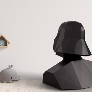 Puede incluir: Un busto negro y geom&eacute;trico de Darth Vader, un personaje de Star Wars, est&aacute; sobre un suelo de madera. El busto est&aacute; hecho de planos angulares y planos. Una estanter&iacute;a con objetos decorativos est&aacute; en el fondo.