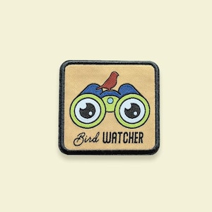 Op de afbeelding: Een geborduurde patch in bruin, beige en blauw met een verrekijker en een kleine rode vogel die er bovenop zit. De tekst "Bird Watcher" is in zwart onder de verrekijker geborduurd.