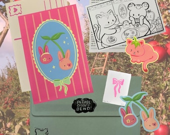 May include: A collection of stationery items including a pink postcard with a blue oval featuring two bunny-cherry hybrids, a black and white coloring sheet with a cat and bear, a pink bow sticker, a strawberry cat sticker, and a green envelope.