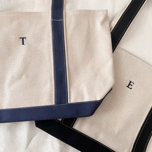 May include: Two canvas tote bags with navy blue and black handles. The bag on the left has the letter 'T' embroidered on the front. The bag on the right has the letter 'E' embroidered on the front.