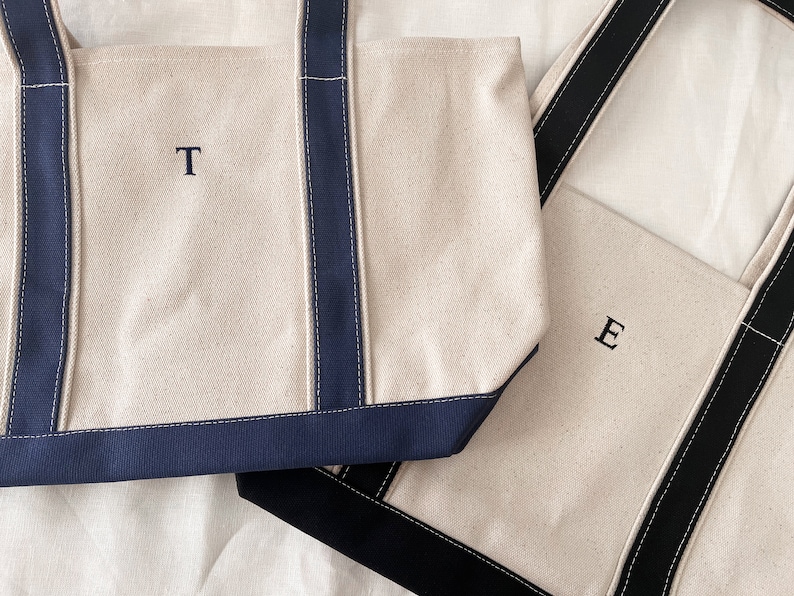 May include: Two canvas tote bags with navy blue and black handles. The bag on the left has the letter 'T' embroidered on the front. The bag on the right has the letter 'E' embroidered on the front.