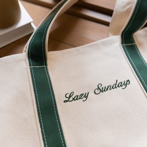 May include: A white canvas tote bag with green trim and the text 'Lazy Sunday' embroidered in green.