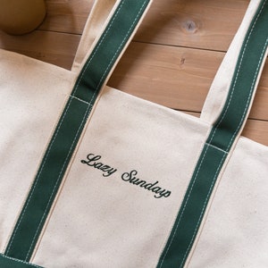 May include: A white canvas tote bag with green straps and the text 'Lazy Sundays' embroidered in black.