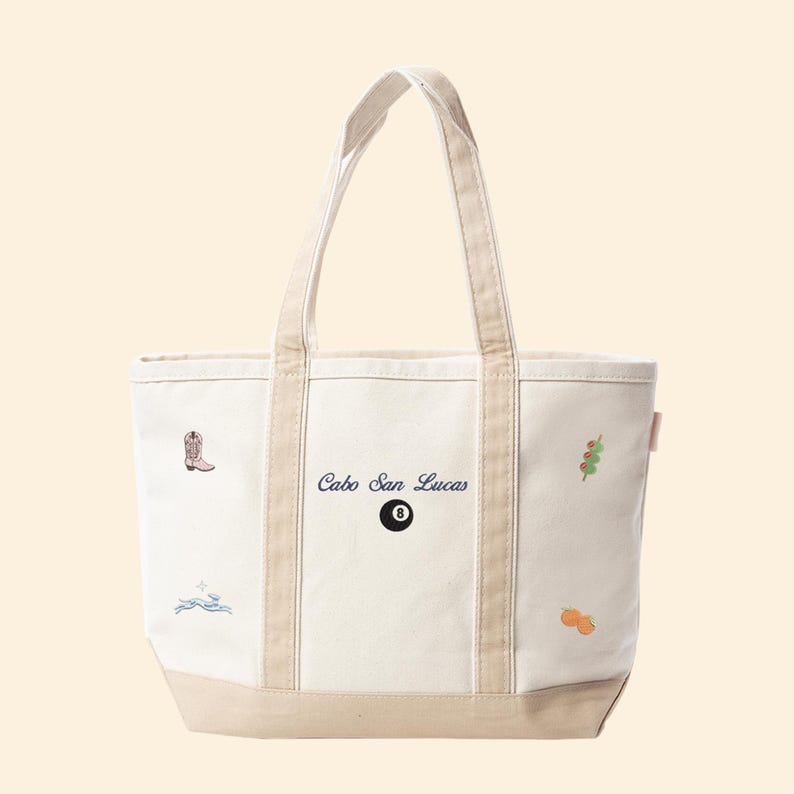 May include: Cream-colored tote bag with beige accents and handles. The bag features embroidered designs including a cowboy boot, a wave, a pool ball with the number 8, and the text "Cabo San Lucas". Additional illustrations include a sprig and an orange.