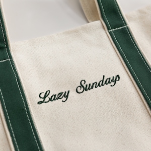 May include: A white canvas tote bag with green fabric trim and the text 'Lazy Sunday' embroidered in green thread.