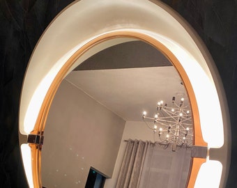 Puede incluir: Un espejo redondo iluminado con un marco dorado y una luz blanca curva. El espejo refleja una habitación con una lámpara de araña, cortinas y papel pintado oscuro. El diseño es moderno y elegante. Diámetro aproximado de 60 cm.