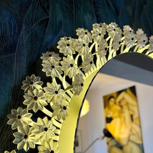 Op de afbeelding: Een ronde spiegel met een gouden lijst en een decoratieve rand van witte plastic bloemen. De spiegel reflecteert een kamer met een bank en een schilderij.