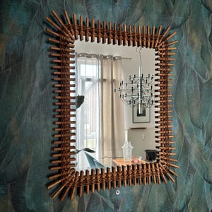 May include: A rectangular wall mirror featuring a brown woven rattan frame with a sunburst-style edge. The frame consists of vertical reed-like spokes radiating outward from the central glass pane, creating a textured, natural appearance.