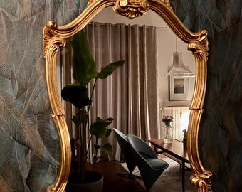 May include: An ornate gold mirror with a shell-shaped top detail and a curved, decorative frame. The reflection shows a room with a plant, chair, and curtains. The mirror is a decorative home decor item.