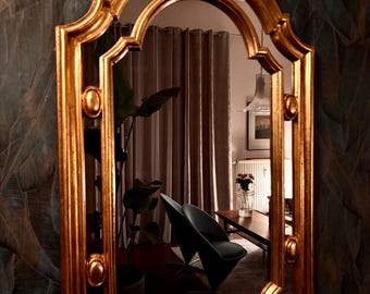 May include: A gold-coloured framed mirror with an arched top and decorative oval accents. The mirror reflects a room with a black chair, a table, and a plant. The frame has a detailed, ornate design.