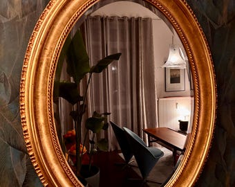 May include: An oval mirror with a gold-coloured frame and a rope-style edge. The mirror reflects a room with a plant, a blue chair, a table, and a lamp. The mirror is mounted on a wall with a dark green and blue leaf pattern.